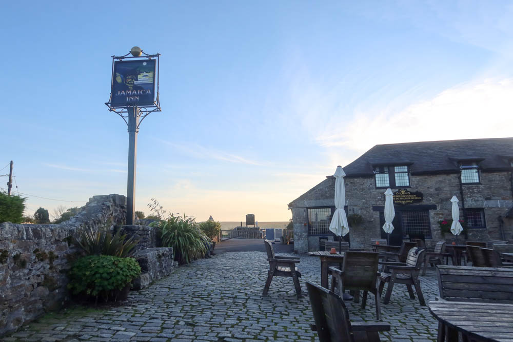 Outside of Jamaica Inn, the sign and stone pub is in the shot. The sunset is in the background and there's a cobblestone courtyard.
