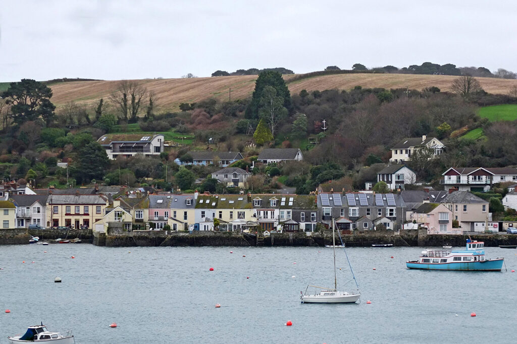 A view from Falmouth over the river to Flushing in Cornwall. There are boats sailing along the river. Houses are lining the water and there are hills and trees in the background. 