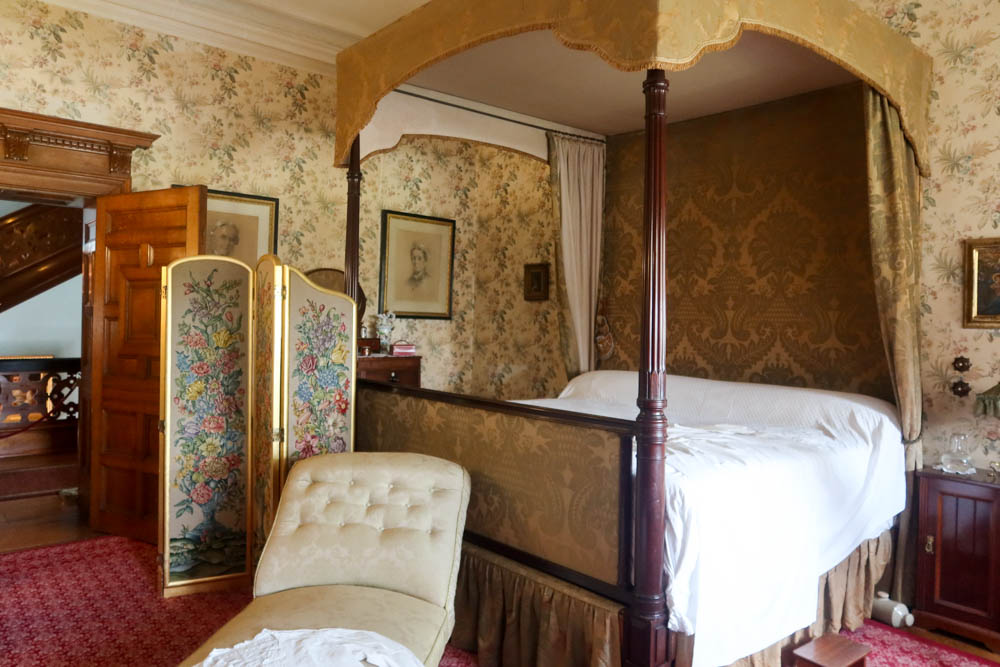 Victorian-style bedroom with a four poster bed with white sheets and a green chair in Lanhydrock, one of the best National Trust properties in Cornwall