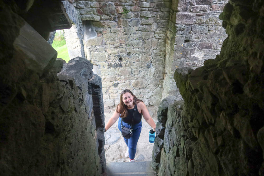 Girl looking up the stairs of lydford castle. She's about to walk up them. She's wearing blue jeans and a black top and is smiling. 