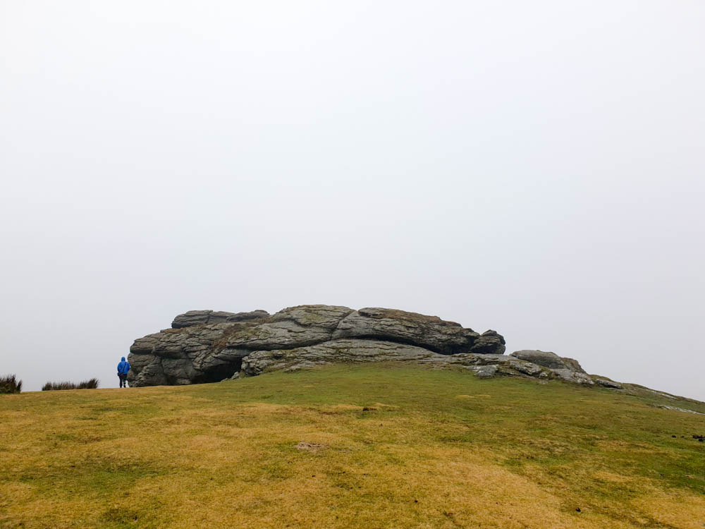 The Rocks of Saddle Tor, with rocks in the middle and grassy landscape underneath. There are clouds in the sky.
