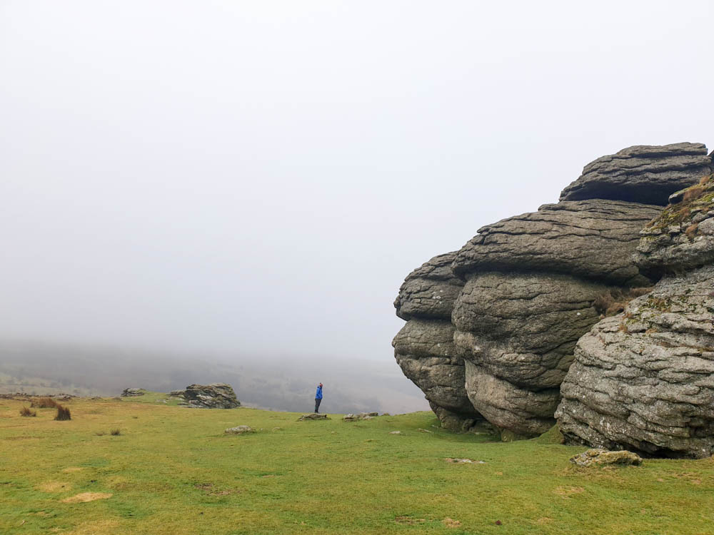 The barren, rocky landscape of Saddle Tor in Dartmoor National Park. There are big rocks on the right and a green landscape on the left. The sky is cloudy.