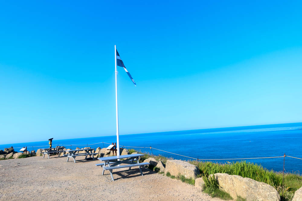 A cornish flag flying at Land's end. It's looking out over the blue sea and the sky is cloudless. 