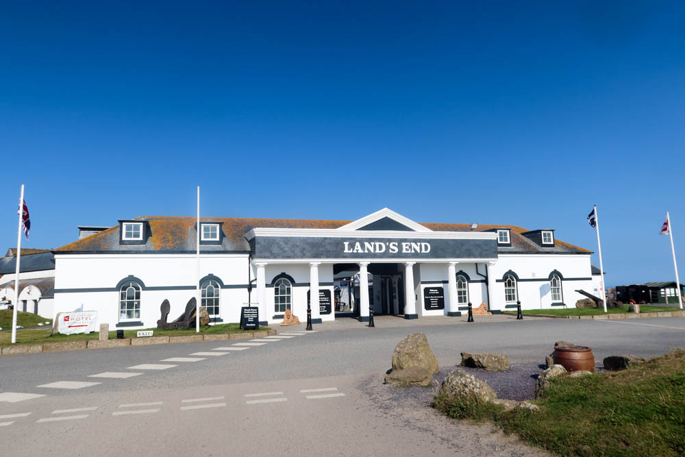 The Land's End landmark attraction building. It's a wide white building with a teal sign and white lettering. The sky behind is cloudless.