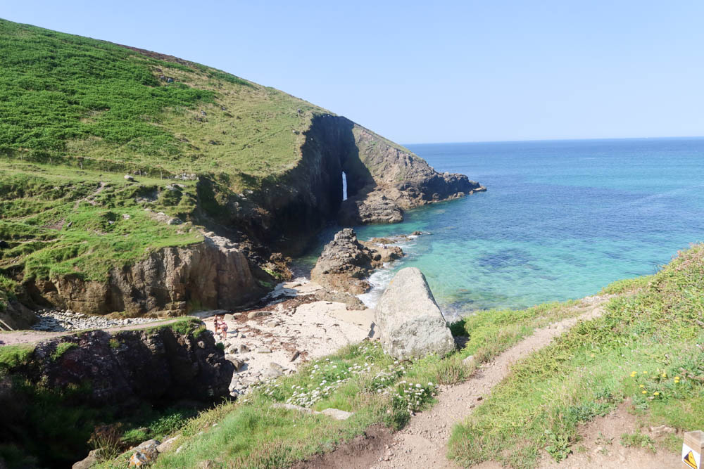 Nanjizal Beach, a beach near Land's End. The photo is from the South West Coast Path. There's sea in the top right corner and a cliff with green grass and a small arch on the other side.