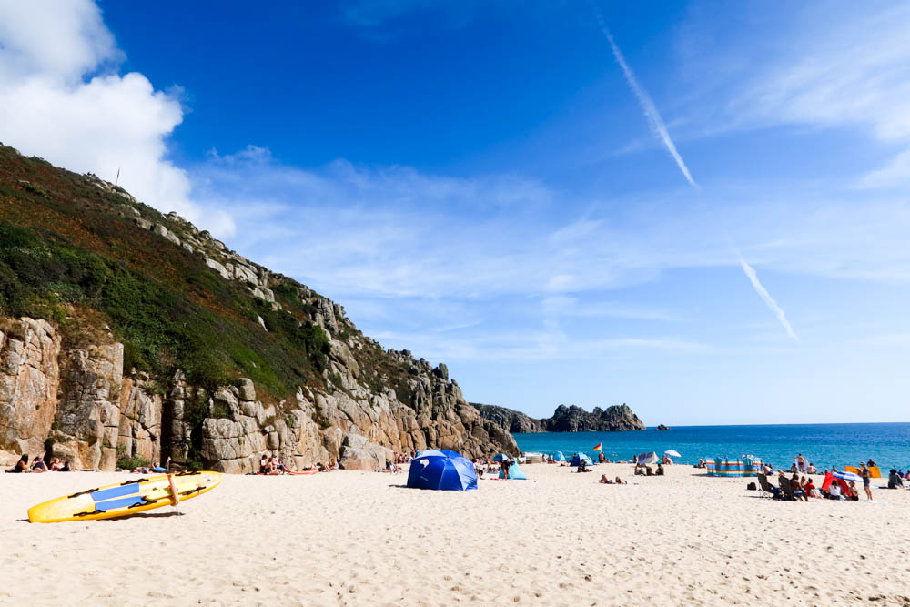 White sand beach near Porthcurno with rocky stacks in the distance and blue water. There's light cloud coverage in the sky.