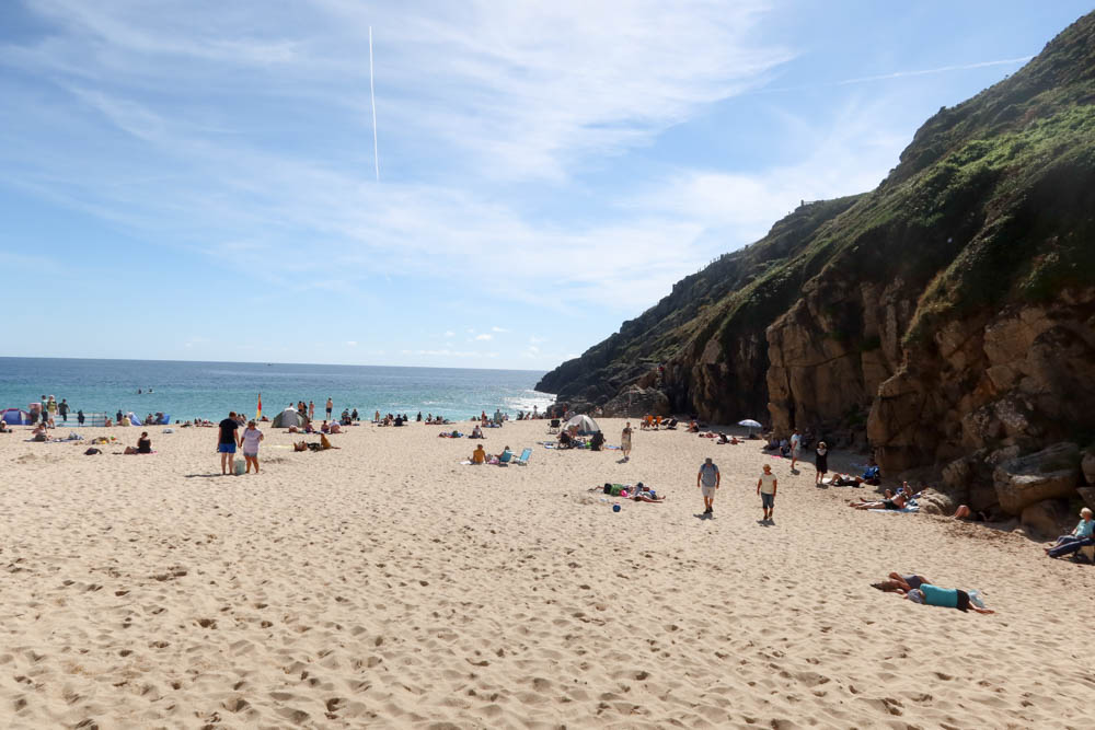 Beautiful Porthcurno Beach with white sound and large looming cliffs, with blue sea in the background.