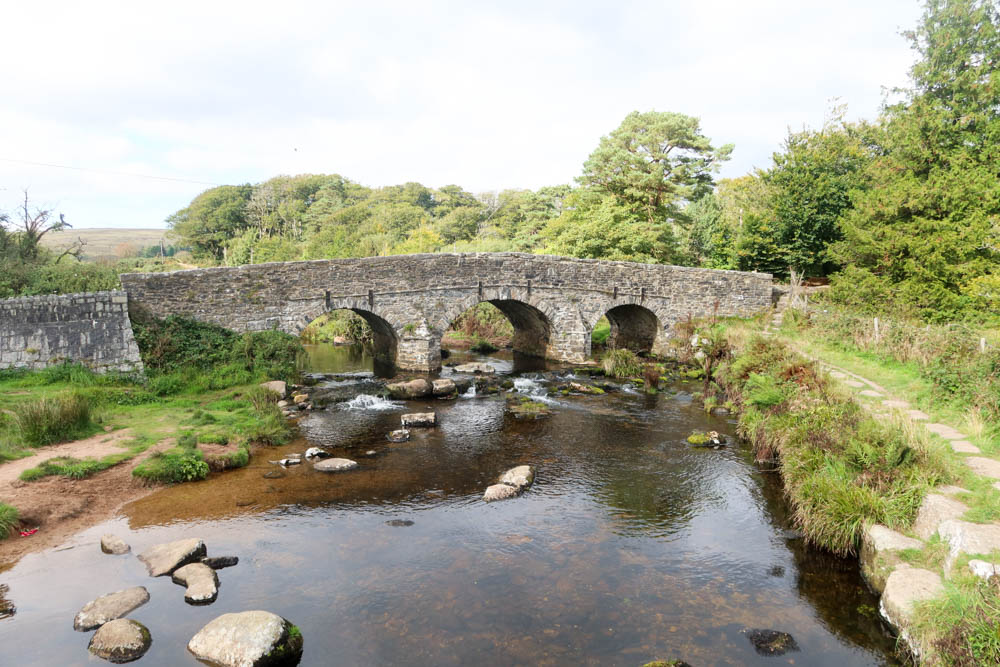 The 18th century bridge at Postbridge in Dartmoor National Park. It has three archways and a river going under the bridge. There are trees in the background and water in the foreground.