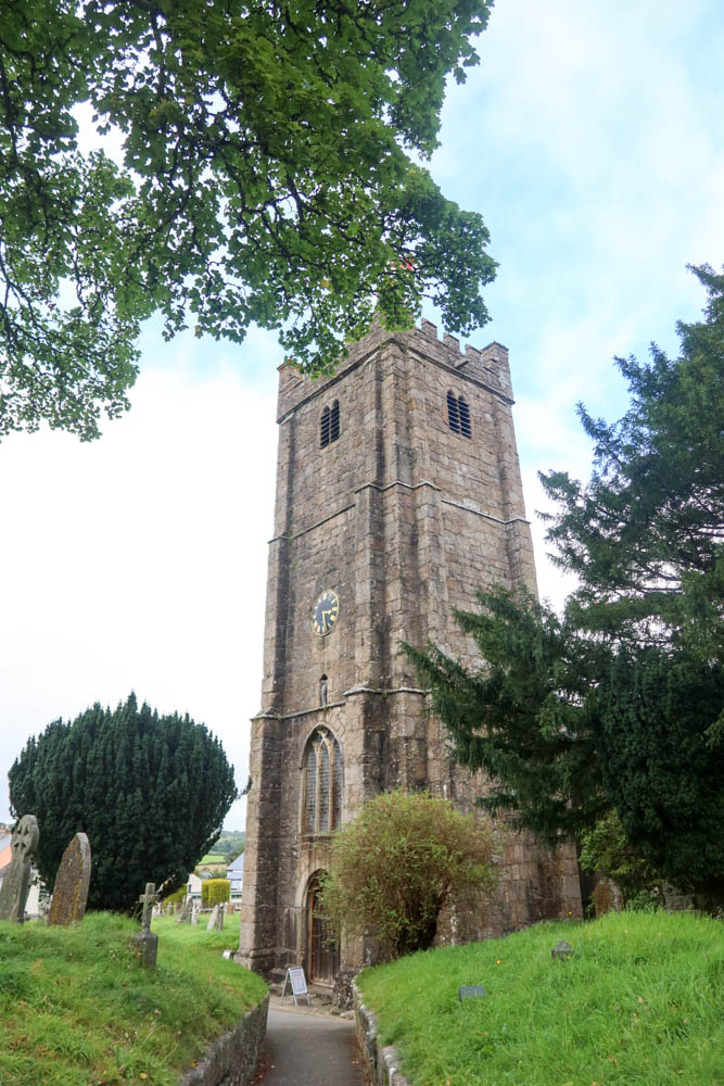 A portrait photo of the church at Chagford, with a large granite tower, green grass in the foreground and blue sky in the background. 