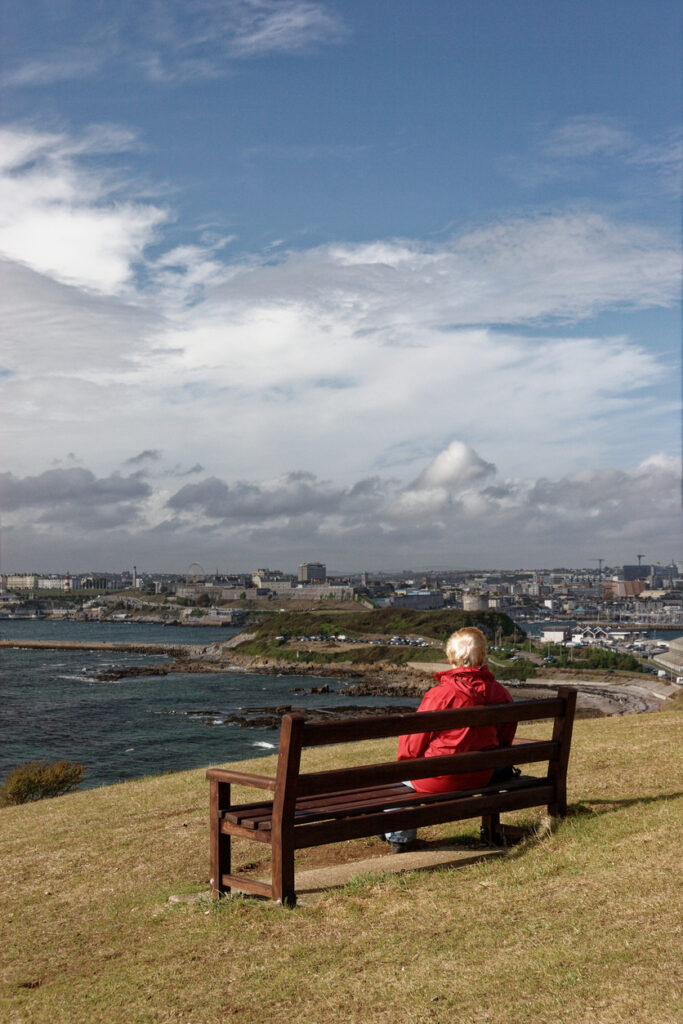 Lady sitting on seat with back to camera looking out over the vista of Plymouth Sound to Drake's Island, Plymouth Hoe and the city. Blue sky light cloud.