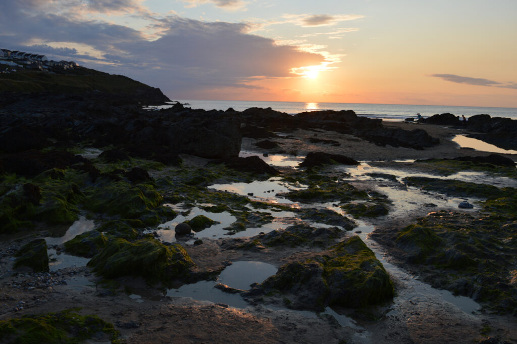 Early evening photo of the rock pools on Fistral beach Newquay, Cornwall, South West England.  The sun is setting and very low in the sky, slightly obscured by clouds.  The tide out exposing the rock pools, sand and rocks.
