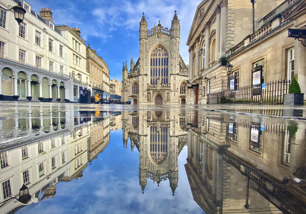 Bath Abbey, The Roman Baths and Pump Rooms in Bath, England pictured below blue skies with fluffy clouds. Recently melted snowfall - and national lockdown - provided the opportunity for stillness and a perfect reflection in a large pool of motionless water.