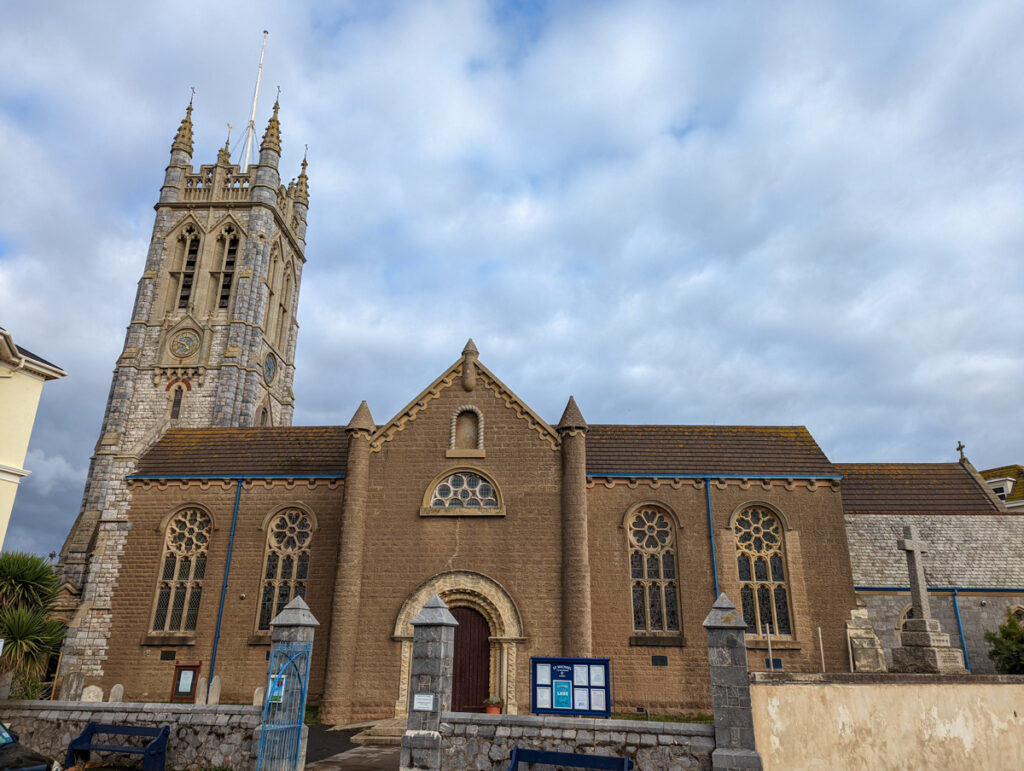 St Michael's Church, which is opposite the sea wall which runs along Teignmouth Beach. The church is a light brown colour and has a tower on the left side.
