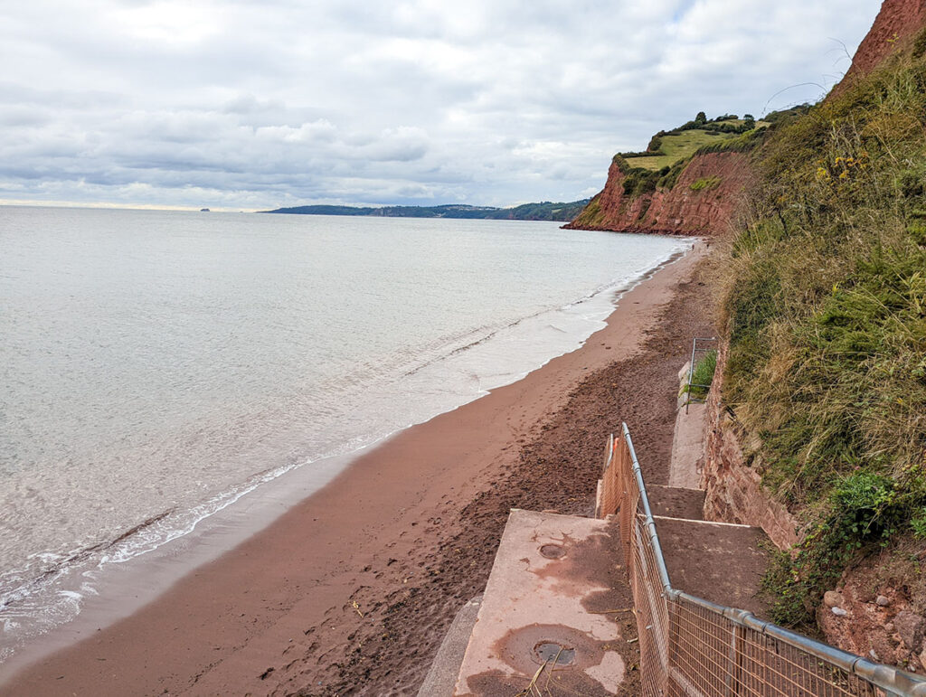 View of Ness Cove Beach, one of the best Teignmouth Beaches with red cliffs and the blue waves on the left.