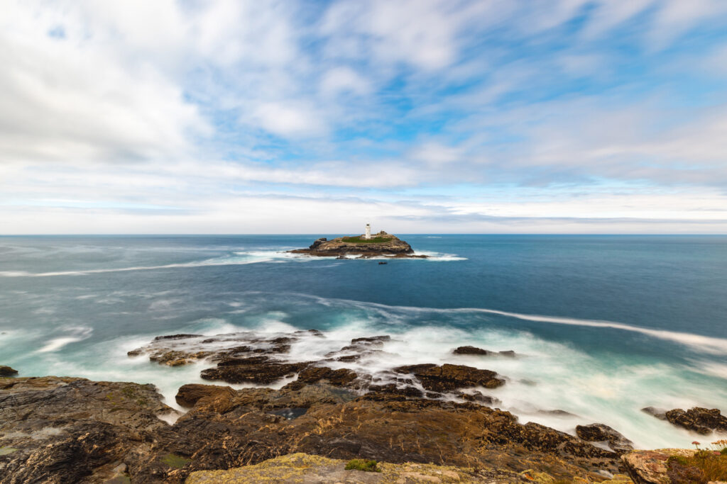 Panoramic long exposure shot of lighthouse at seaside in Cornwall at Godrevy point with waves and rocks on foreground and blue cloudy sky on a summer day - Travel and landscape