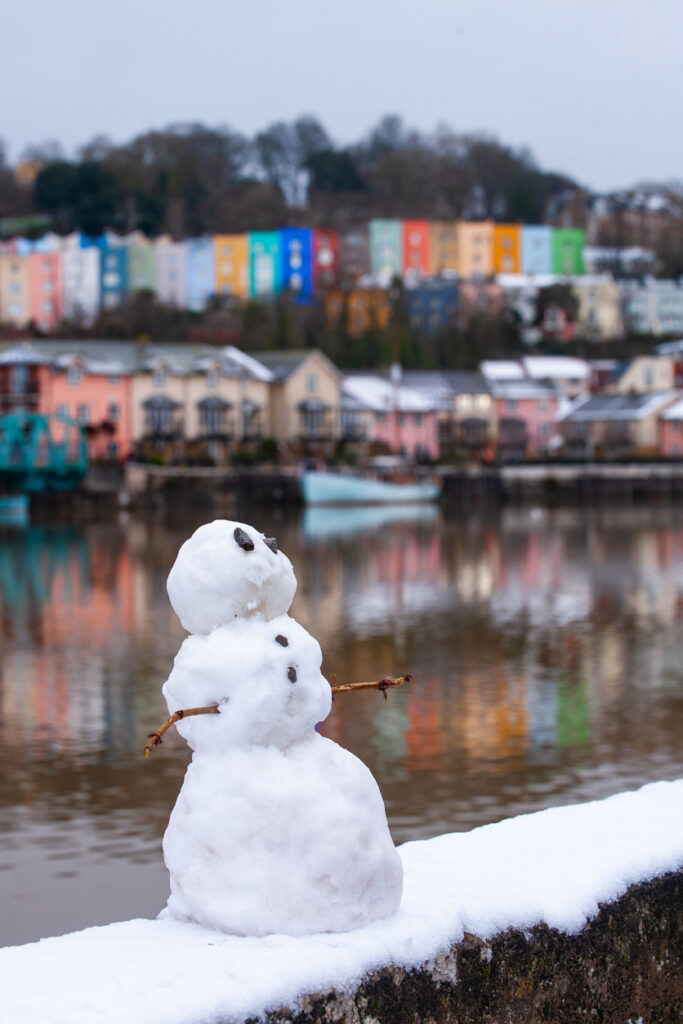 A small snowman on the ground at Bristol Harbourside. There are colourful houses out of focus behind it, which are also reflected in the river.
