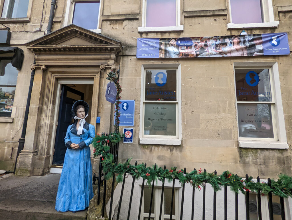 A statue of Jane Austen, wearing a blue dress, outside of a Georgian building with houses the Jane Austen museum in Bath.