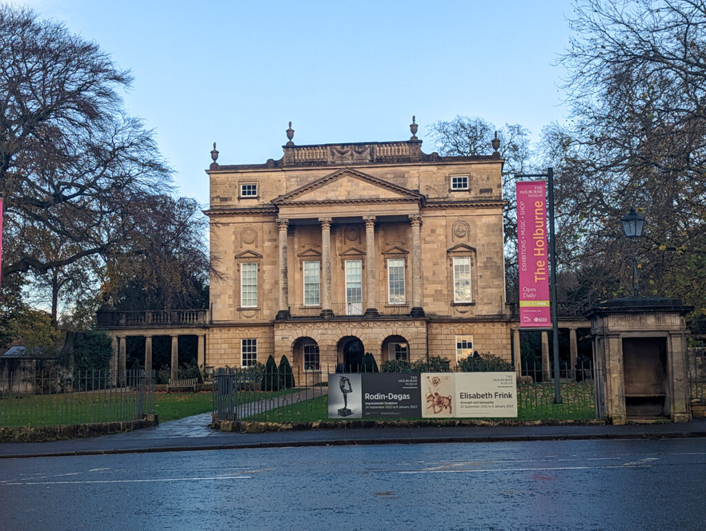The Holburne Museum, which is a Georgian building in Bath, there are trees on either side and the road in the foreground.