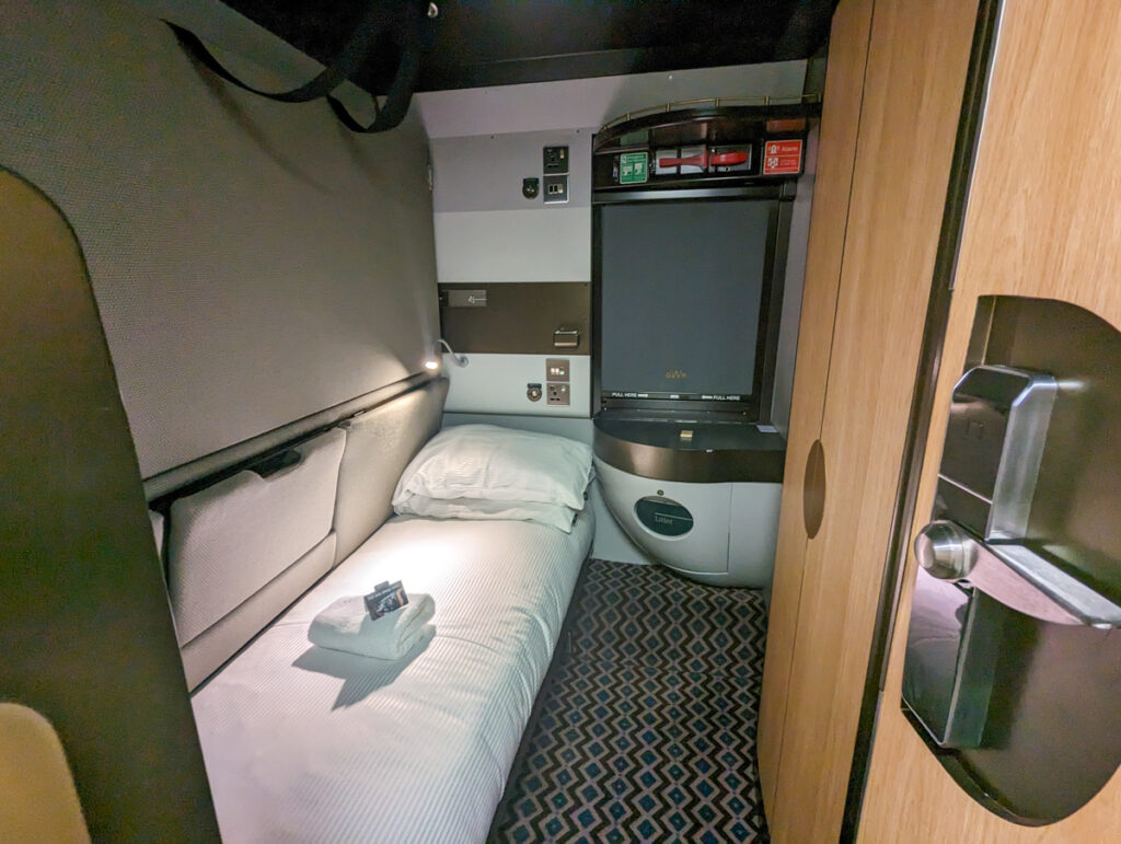 The Night Riviera Sleeper cabin, with the doorfame in the foreground and a bed with a towel, bedside table that folds up to be a sink, rubbish bin, blackout blind
