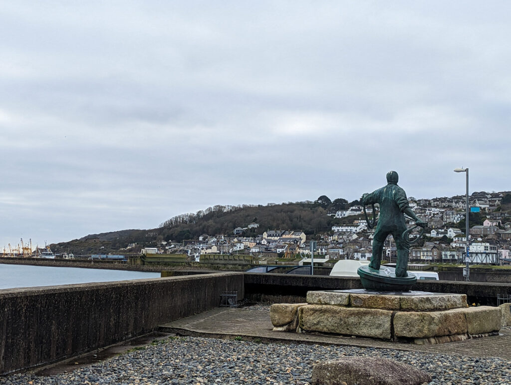 A statue in the foreground with the buildings of Newlyn in the background, with the sea and grey skies