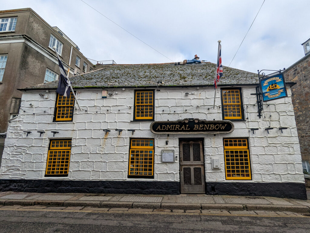 The Admiral Benbow Inn which is a historic pub with smuggling connections in Penzance. It's a white building with yellow windowframes and a pirate figure on the roof.