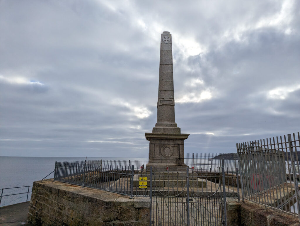 War memorial in Penzance, dedicated to all of the men from Penwith that fought in the World Wars. It stands against the sea and a cloudy sky. 