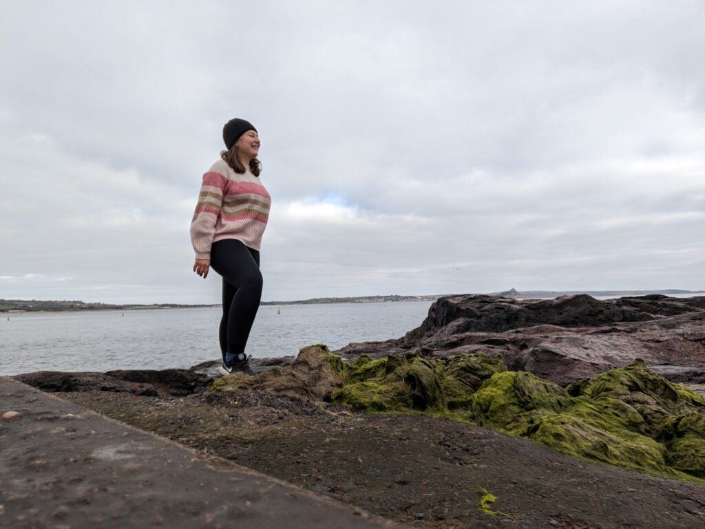 Girl standing on Battery Rocks in Penzance, wearing a pink jumper and black leggings, looking into the distance.