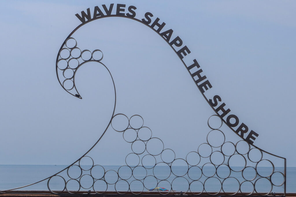 Sculpture stating "waves shape the shore" with blue sky in the background.