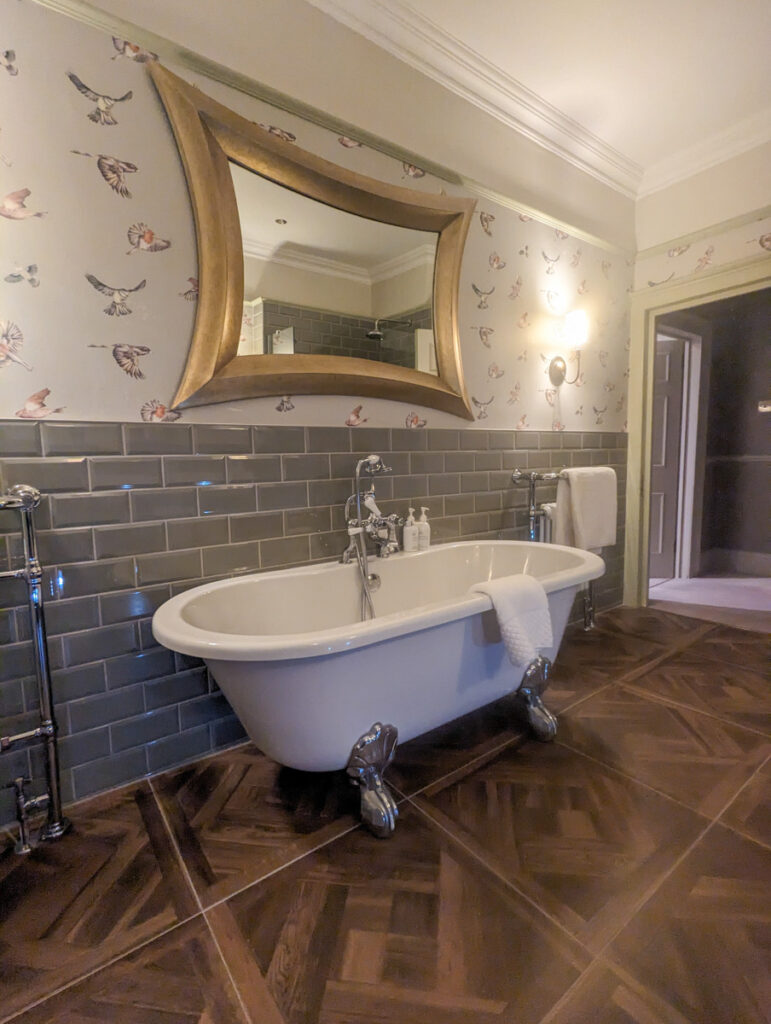 Bathroom with vintage charm, showcasing a freestanding clawfoot bathtub, olive green subway tiles, parquet flooring, and bird print wallpaper