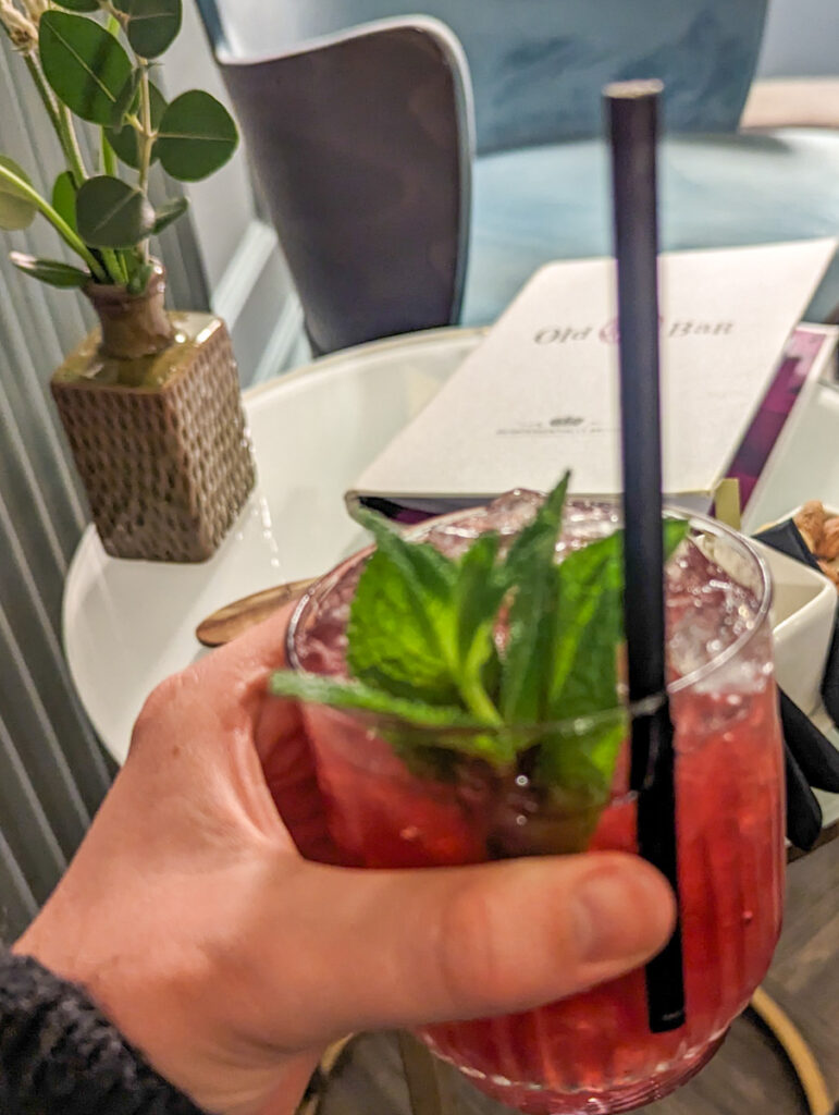 A close-up of a hand holding a refreshing cocktail garnished with fresh mint leaves, in a dimly lit lounge with blurred decor in the background.