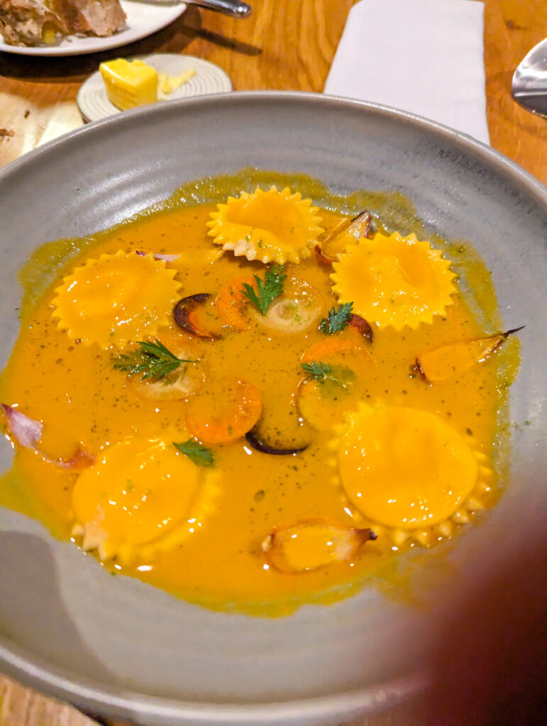 A colorful plate of ravioli in a butternut squash broth with carrot and herb garnish, presented on a rustic grey plate, ready for a gourmet dining experience.