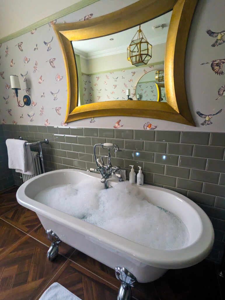 A luxurious bathroom featuring a clawfoot bathtub filled with bubbles, set against bird patterned wallpaper and olive subway tiles, reflecting a hint of opulence.