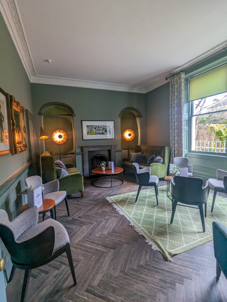 Hotel lounge area with green and grey decor, herringbone wood flooring, and elegant seating arrangements under a traditional fireplace mantle.