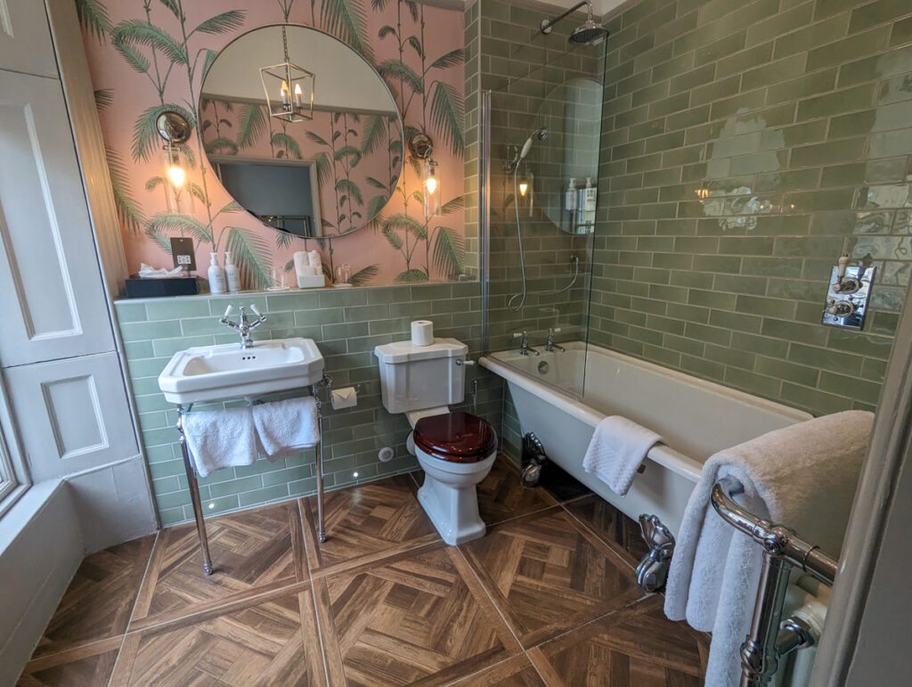 Elegant bathroom with pastel pink wallpaper featuring palm motifs, olive green subway tiles, a classic white bathtub, pedestal sink, and vintage-style fixtures.
