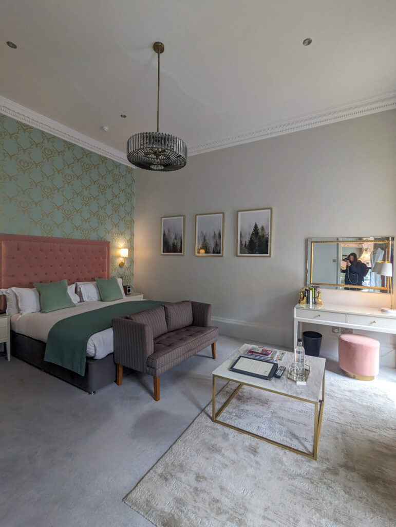 Chic hotel room with a plush king-sized bed, pink headboard, patterned wallpaper, chandelier lighting, and a cozy sitting area with a gray sofa.