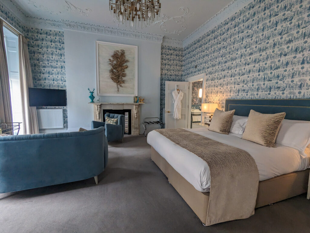 Luxurious hotel bedroom with blue velvet sofa, white and beige bedding, feather artwork over a marble fireplace, and patterned wallpaper creating a tranquil atmosphere.