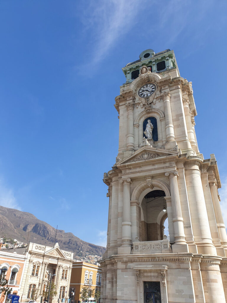 A majestic clock tower stands against a clear blue sky, featuring intricate baroque architecture with statues and a visible clock face, surrounded by the mountainous landscape of Pachuca, Mexico.