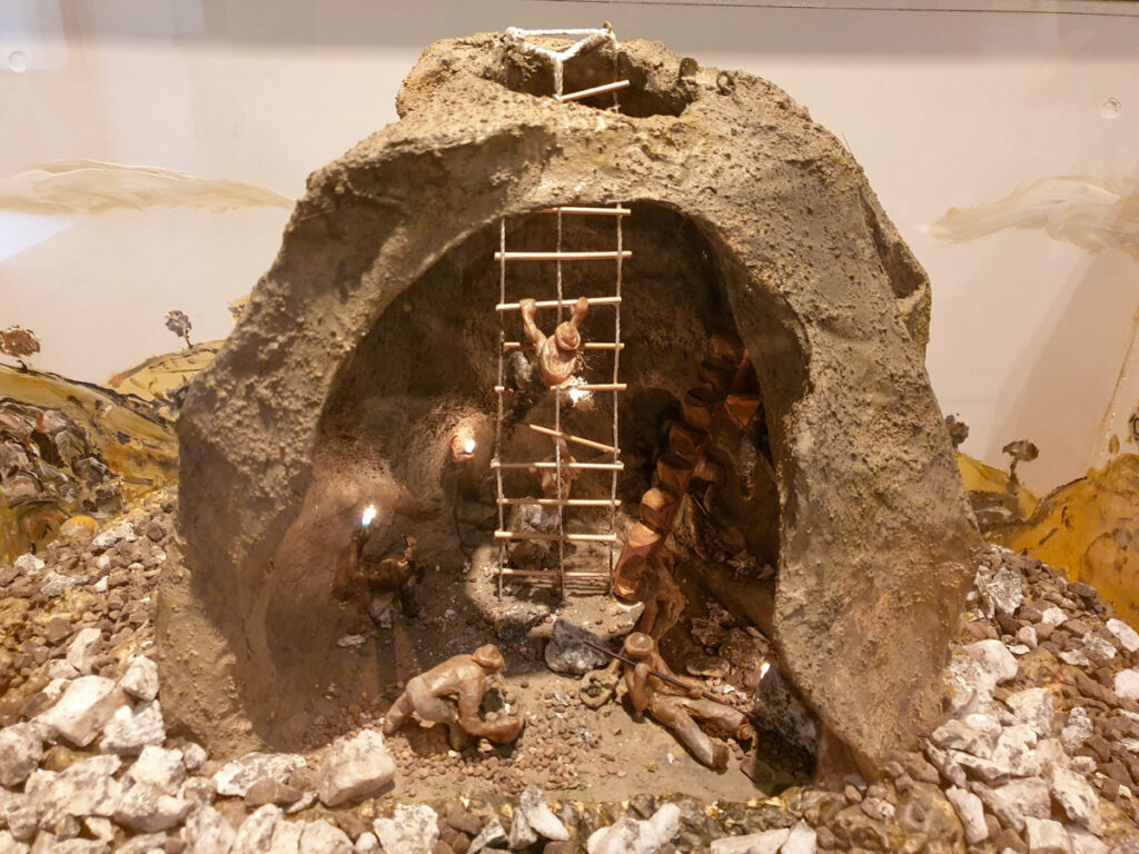 Detailed model display of a historical underground mine with miniature figures of miners climbing ladders inside a rocky structure, exhibited in a museum.