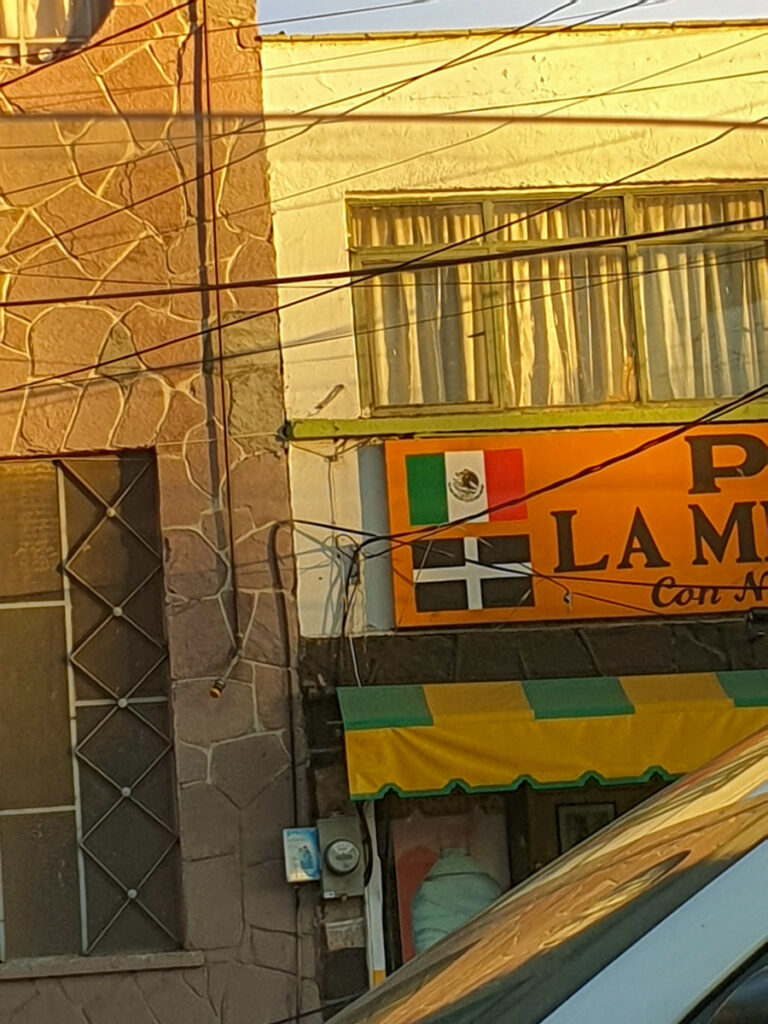 Sunlit facade of a rustic building displaying a colorful banner with the flags of Mexico and Cornwall, representing the cultural connection in a mining town.