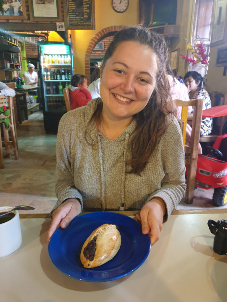 A smiling woman sitting inside a cozy café, holding a freshly baked pasty on a blue plate, with a casual and inviting atmosphere.