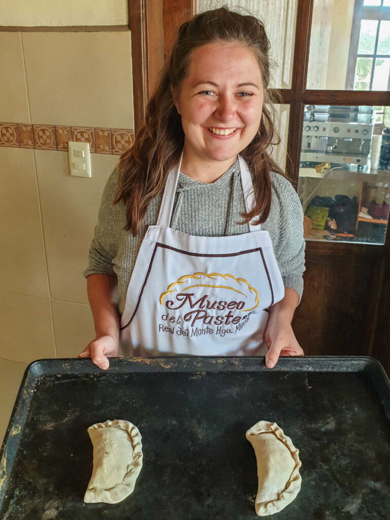 A woman in a cooking apron joyfully prepares traditional pasties, standing in a kitchen with two raw pasties on a baking tray in front of her