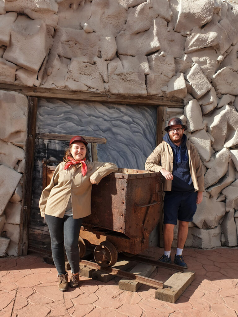 "A couple dressed in casual outdoor attire posing with an old mining cart at a historic mining site, surrounded by walls made of white sacks.