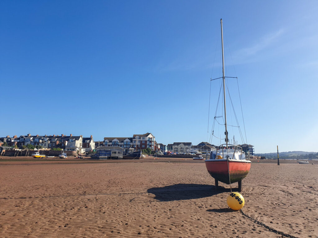 A sailboat with a red hull is anchored on a sandy beach under a clear blue sky. In the background, there are houses lined up along the shoreline.