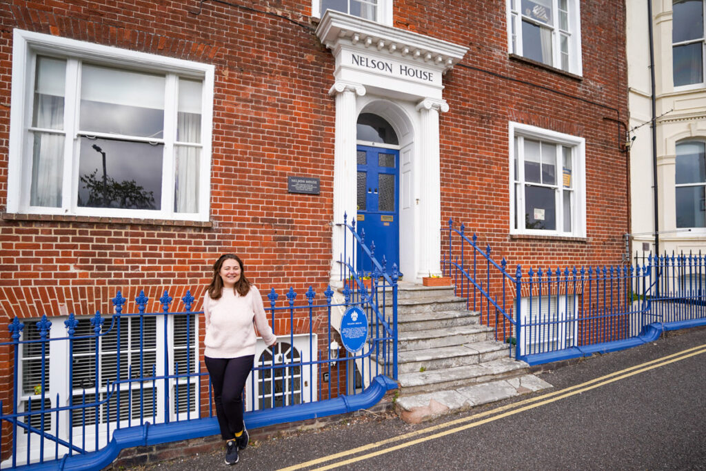 A woman in a light sweater and dark pants stands in front of a brick building named Nelson House, which has a blue door and white columns.