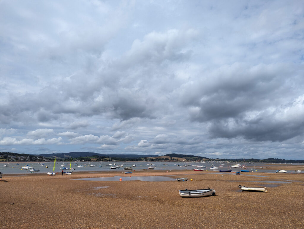 A beach scene under a cloudy sky, with several small boats scattered along the shore and more boats visible in the water, with hills in the distance.