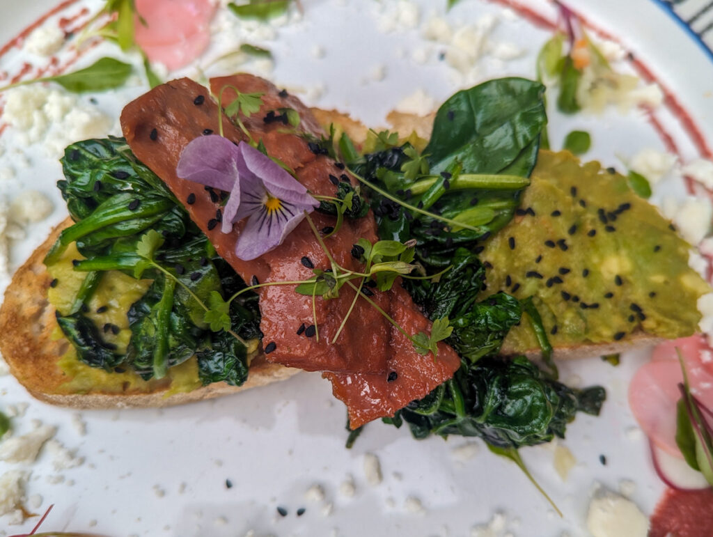 A close-up of an artistic dish featuring avocado toast topped with greens, a slice of plant-based meat, and a small edible flower, presented on a colorful plate.
