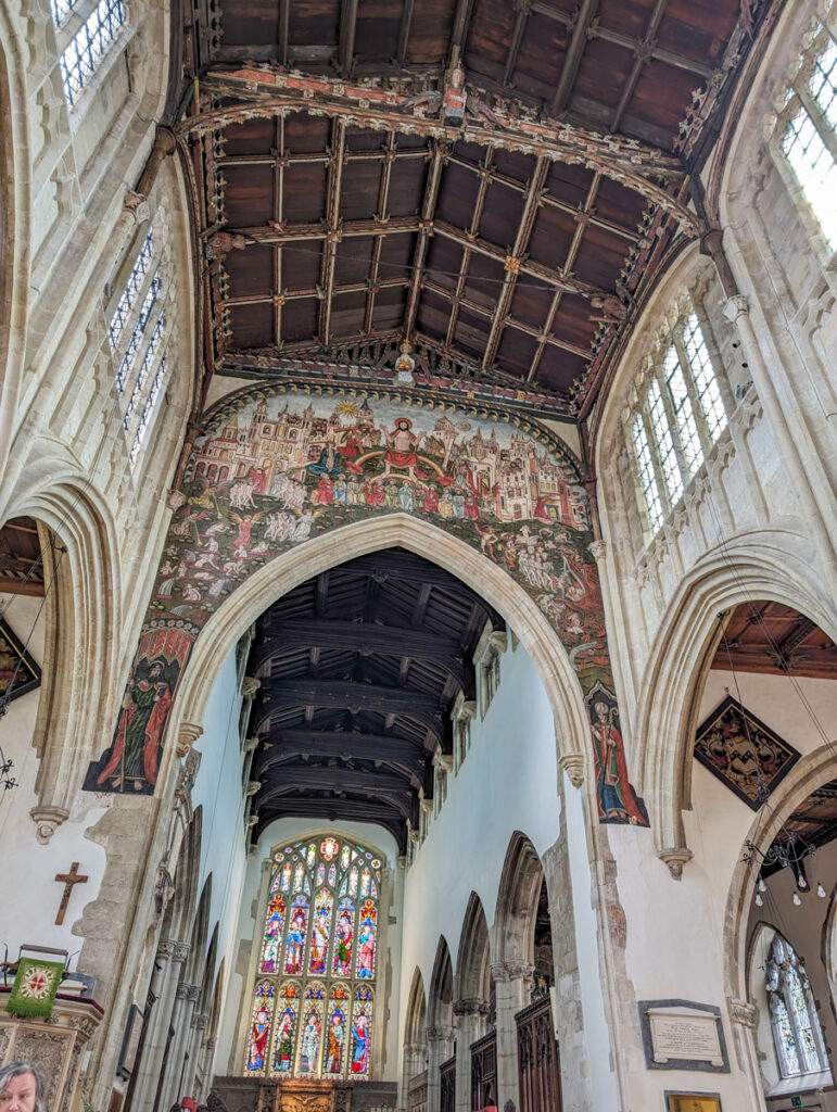 The interior of a Gothic cathedral with an ornate painted ceiling depicting religious scenes, towering arches, and stained glass windows in the background. The scene captures the grandeur and historical artistry of the church's architecture.