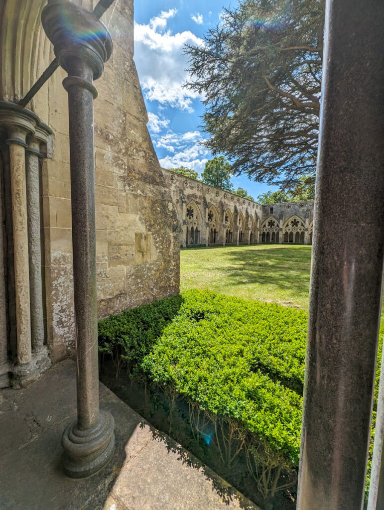 A view from within the cloisters of Salisbury Cathedral, looking out onto a well-maintained courtyard with green hedges and a large tree, framed by Gothic arches and stone columns.