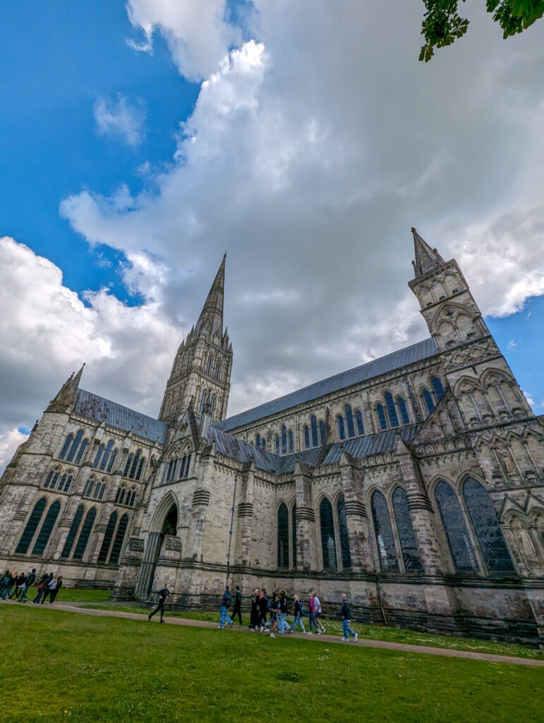 The exterior of Salisbury Cathedral with its tall, pointed spire reaching into a partly cloudy sky. The large, stone structure is seen from a low angle, emphasizing its Gothic architecture and the expansive lawn in front of it.