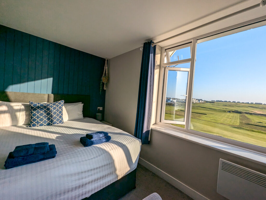 A cozy bedroom featuring a neatly made bed with blue towels and a patterned cushion, against a dark accent wall. Sunlight streams through a large open window offering a scenic view of green fields and clear blue skies.